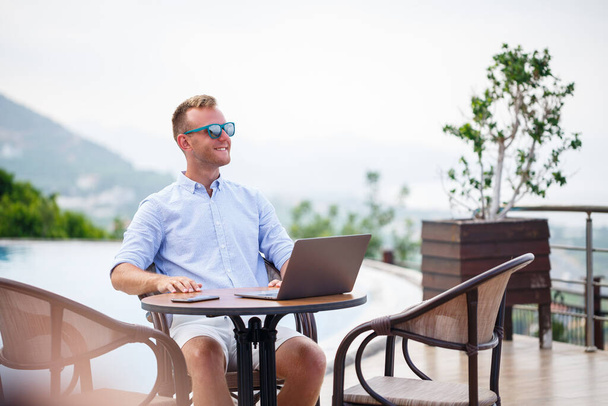 サングラスの成功したハンサムな男性実業家は、プールの近くに座ってラップトップで動作します。遠隔作業だ。フリーランス - 写真・画像