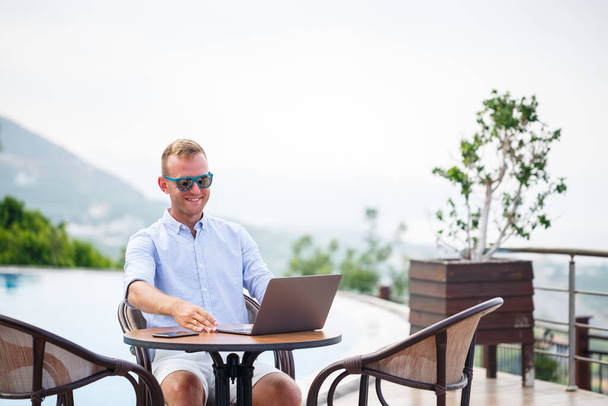 El exitoso hombre de negocios guapo con gafas de sol trabaja en un portátil sentado cerca de la piscina. Trabajo remoto. Autónomo - Foto, imagen