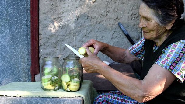 Ζαρωμένη ηλικιωμένη γυναίκα χαμογελάει και κόβει ωμά κολοκυθάκια και τα βάζει σε βάζο για σπιτική κονσερβοποιία με σπόρους άνηθου και σκόρδο. Αγροτική τοπική κουζίνα με βιολογικά λαχανικά - Φωτογραφία, εικόνα