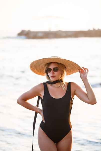 Beautiful girl in a black swimsuit and hat on a sandy beach at the sea in the sunset sunlight - Photo, Image