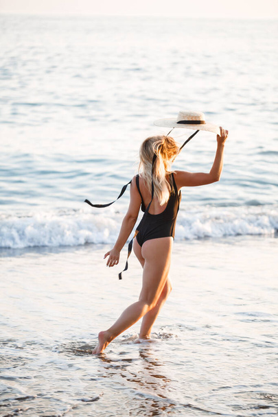 Hermosa chica en un traje de baño negro y sombrero en una playa de arena en el mar en la luz del sol puesta del sol - Foto, Imagen