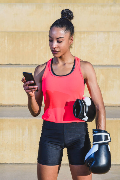 vertical portrait of an attractive afro american woman dressing sport clothes and holding black and white boxing gloves using her smart phone while standing in a park - Фото, изображение