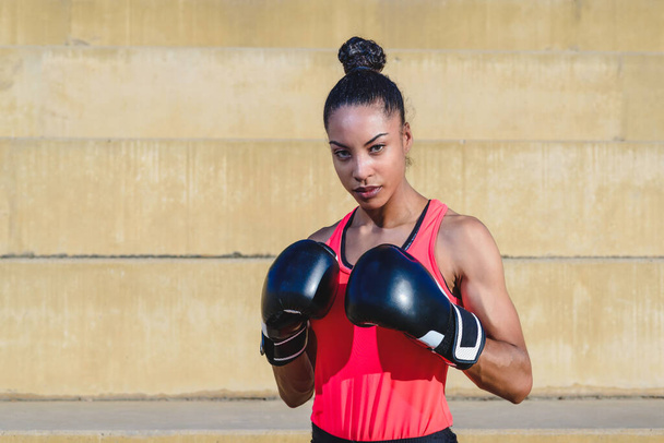 horizontal portrait of an attractive african woman wearing sport clothes and boxing gloves standing in defense pose - Foto, Imagem