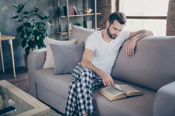 Portrait of his he nice attractive focused bearded guy sitting on divan reading interesting paper book page print spending free time leisure at modern loft industrial interior style indoors - Foto, Imagem