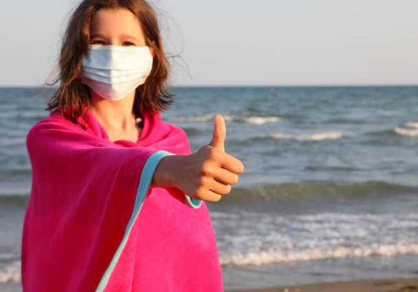 petite fille avec le masque chirurgical ne Ok tenant le pouce de la main levée par la mer en été - Photo, image