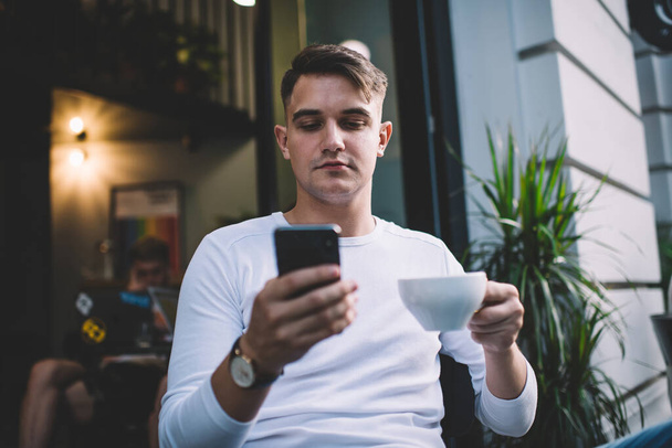 Koncentrált fiatalember visel alkalmi ruhát, és válaszol e-mail a mobiltelefon alatt kávészünet a nyári időszakban a modern kávézó területén - Fotó, kép