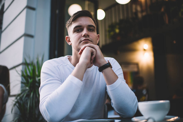 Low angle of pensive young male in casual clothes and wristwatch sitting at table in cafe and looking at camera - Foto, Imagem