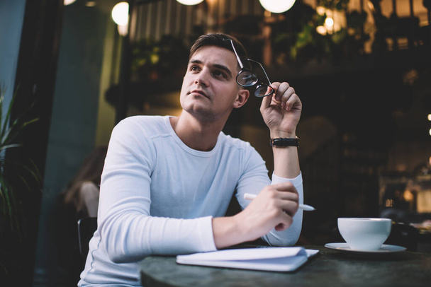 From below of thoughtful male student in casual outfit and wristwatch pondering about essay while sitting at table in cafe with glasses in hand and looking away - Φωτογραφία, εικόνα
