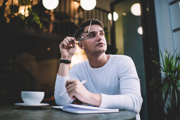 Low angle of thoughtful young author in casual clothes working on book while sitting at table in cafe with eyeglasses in hand and looking away - Фото, изображение