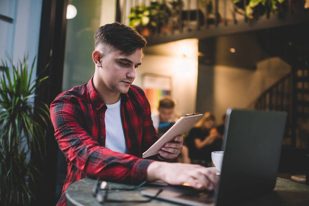 Zdola zamyšlený mladý muž v neformální košili hledá informace ze schránky na notebooku, zatímco sedí u stolu v kavárně - Fotografie, Obrázek
