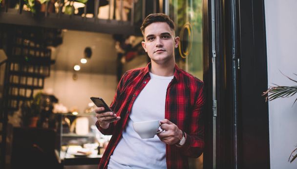 Low angle of cheerful male wearing casual clothes and browsing smartphone while standing near entrance of modern cafe and looking at camera - Foto, Bild