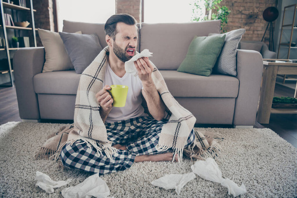 Full length photo of sick ill guy sit carpet hold hot tea beverage mug sneeze napkin cover plaid checkered blanket wear nightwear sleepwear in house indoors - 写真・画像