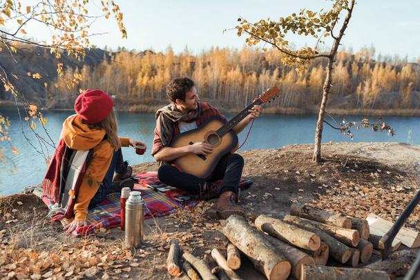 Een koppel rust in de natuur, tegen de achtergrond van de herfstbomen en de rivier. Guy speelt gitaar. - Foto, afbeelding