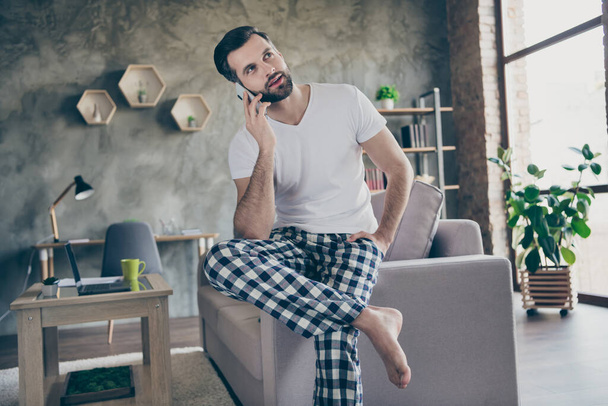 Photo of handsome man sit divan quarantine home call friend smartphone tell say talk speak lifestyle daily news wear checkered plaid pants trousers white t-shirt in house indoors - Foto, immagini