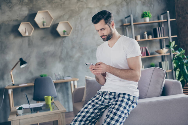 Photo of concentrated man sit divan use smartphone search coronavirus information wear checkered plaid pants trousers white t-shirt in living room house indoors - 写真・画像