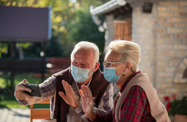 Старшая пара в защитных масках для лица с помощью мобильного телефона для видеосвязи с родственниками во время пандемии и карантина - Фото, изображение