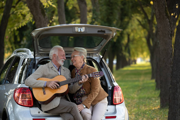 Романтическая старшая пара, сидящая на заднем сиденье машины и играющая на гитаре в лесу - Фото, изображение