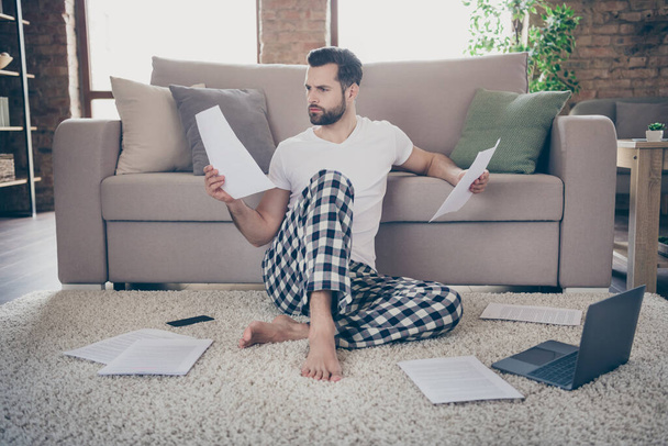 Photo of handsome focused serious guy sit floor sofa work morning browsing notebook freelancer remote work home quarantine read contract look for mistakes pajama living room indoors - Foto, immagini