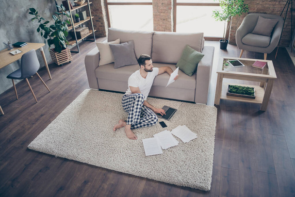 Nice attractive focused guy sitting on carpet studying learning working remotely online analyzing documents at modern loft industrial open space interior style flat indoors - Zdjęcie, obraz