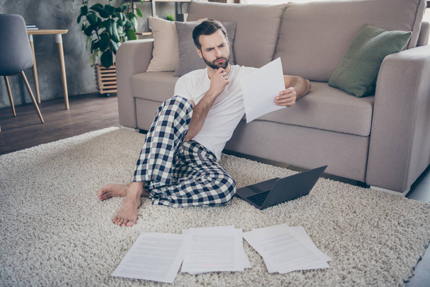 Photo of handsome homey guy sit comfy floor sofa working morning browsing notebook freelancer remote work stay home quarantine read contract think sign or not pajama living room indoors - Фото, изображение
