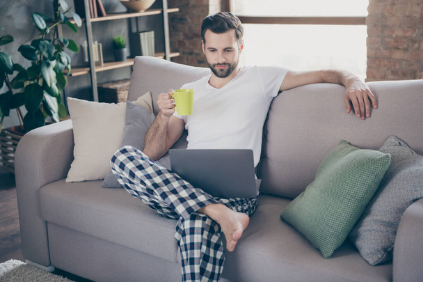 Photo of handsome homey guy sit comfy sofa saturday morning drink fresh coffee browsing notebook freelancer remote work stay home quarantine pajama weekend living room indoors - Foto, immagini
