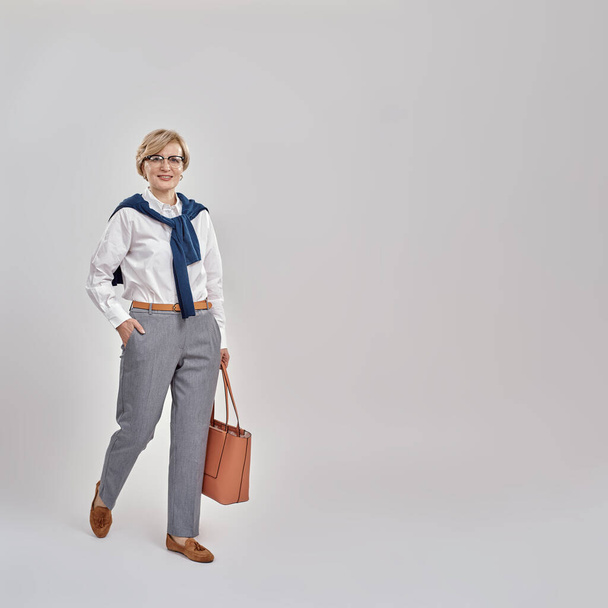 Full length shot of elegant middle aged caucasian woman wearing business attire and glasses smiling at camera while posing, standing with handbag isolated over grey background - Photo, Image