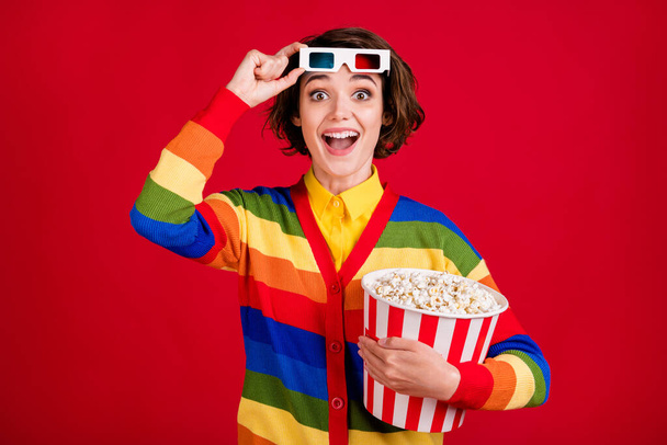 Photo of attractive funny lady hold big popcorn bucket watch movie cinema premiere meet friends take off 3d specs wear striped cardigan jumper isolated bright red color background - Фото, зображення