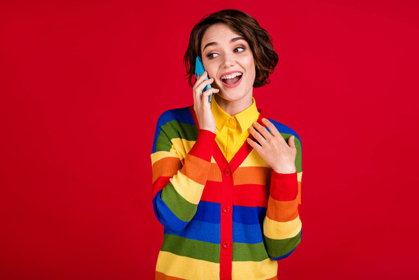 Photo of beautiful excited funny lady hold telephone talking girlfriend best friend listen news sharing fresh gossips rumors wear stripes jumper cardigan isolated red color background - Foto, imagen