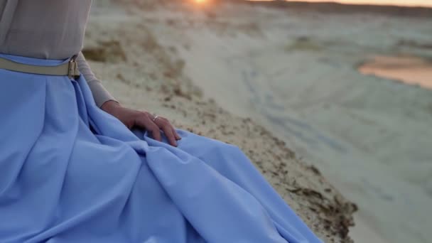 Nahaufnahme einer jungen Brünetten in blauem Rock sitzt auf einem Sandhügel mit Sonnenuntergang im Hintergrund - Filmmaterial, Video