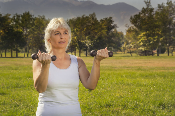 Пожилая женщина поднимает гантели во время занятий фитнесом в городском парке на фоне гор в солнечный день - Фото, изображение