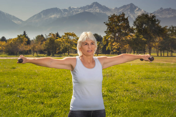 Elderly woman is engaged in fitness with a dumbbell in isolation mode outdoors in a park against a background of mountains on a sunny day - Foto, Bild