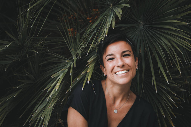 Πορτρέτο μιας όμορφης χαρούμενης γυναίκας κοιτάζοντας την κάμερα χαμογελώντας με τα φυτά στο παρασκήνιο - Φωτογραφία, εικόνα