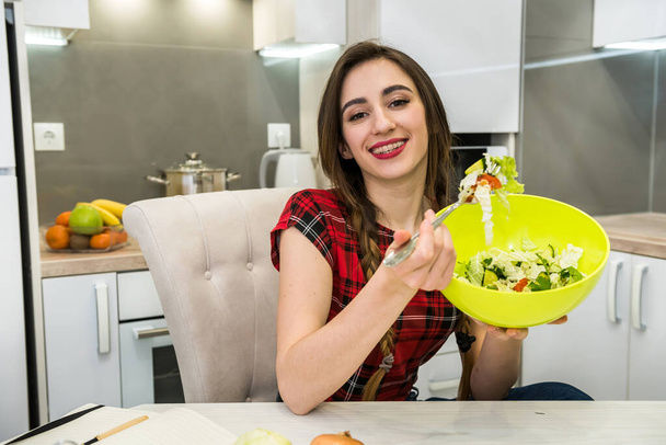 Pretty girl eating salad of fresh vegetables while smiling and sitting in the kitchen at home. - Fotografie, Obrázek