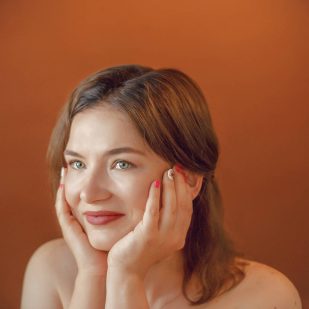 large portrait of a European girl with hands on a brown background. One girl smiles on a plain background. 30 years old. Face. Hand.look, eyes. Face on your hands. - Photo, image