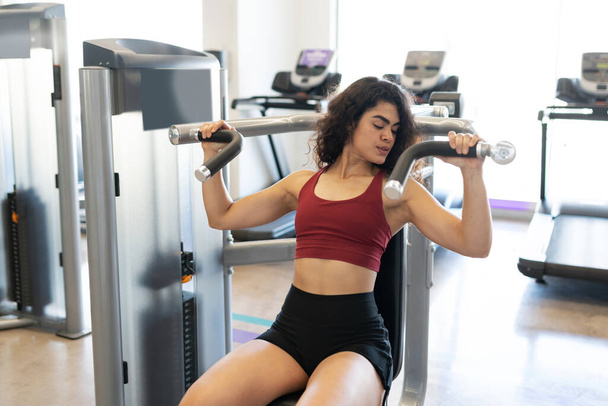 Привлекательная женщина, смотрящая на свои мышцы, сидя в тренажере в фитнес-клубе - Фото, изображение