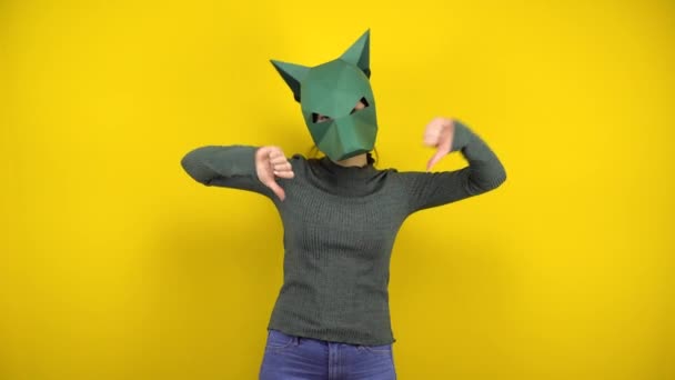 Mulher nova em uma máscara de chacal de papelão mostra desgostos com polegares em um fundo amarelo. Mulher de camisola verde e máscara. - Filmagem, Vídeo