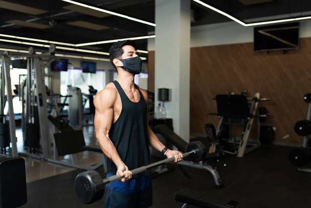 schöner junger Mann mit Gesichtsmaske beim Krafttraining mit der Langhantel im Fitnesscenter - Foto, Bild