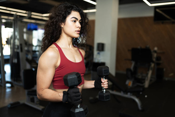 Mujer joven latina con músculos tonificados levantando dos pesos para fortalecer sus bíceps en el gimnasio - Foto, imagen