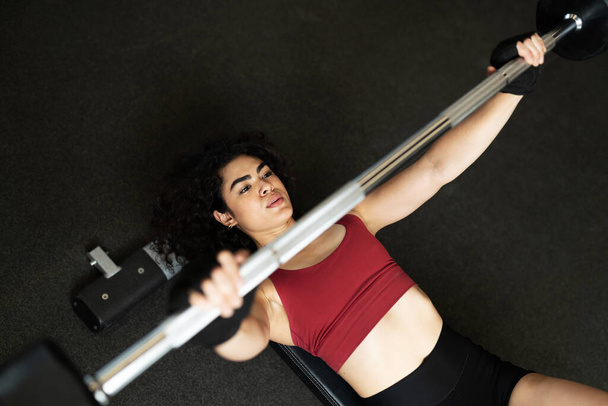Giovane donna ispanica vista dall'alto sdraiata su una panchina e che fa un allenamento di sollevamento pesi con un bilanciere in palestra  - Foto, immagini