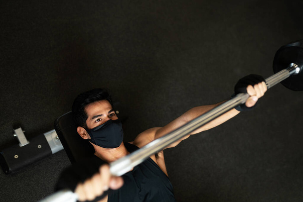 Ângulo alto de um jovem hispânico usando um treinamento de máscara facial com pesos em uma barra no clube de fitness durante a pandemia - Foto, Imagem