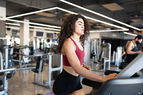 Güzel, kıvırcık saçlı İspanyol kadın modern bir spor salonundaki elektronik koşu bandında antrenmanı bitiriyor. - Fotoğraf, Görsel