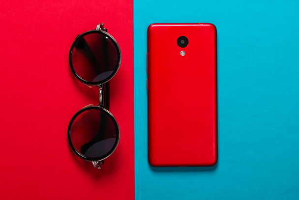 Las gafas a la moda y el smartphone sobre el fondo rojo azul. Accesorios mujer - Foto, Imagen