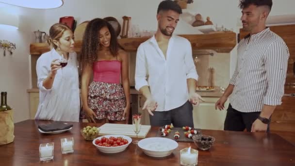 Two young adult mixed race couples having fun in the kitchen preparing the aperitif snacks on the table - Handsome guy making vegetarian skewers while his friends make fun of him joking and laughing - Metraje, vídeo