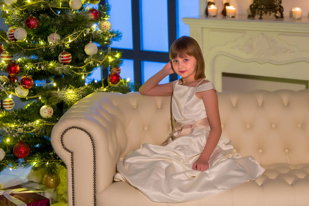 Beautiful little girl sitting on a sofa near the New Year tree. - Foto, imagen