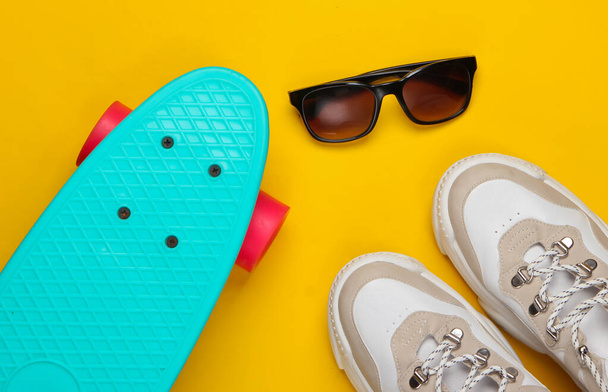 Modieuze sneakers met cruiser board, zonnebril op gele achtergrond. Jeugdplezier. Bovenaanzicht. Minimalisme - Foto, afbeelding
