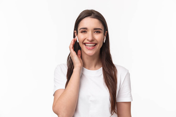 Close-up portrait of attractive smiling happy woman, touching wireless headphone, using earphones to contact friend, having phone conversation with earbuds, laughing carefree - Foto, afbeelding