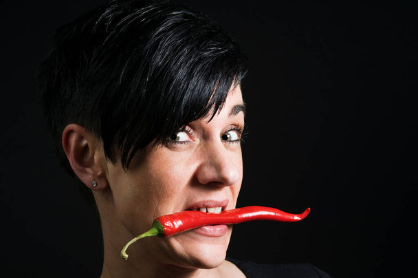 Lateral head portrait of a young black-haired woman in front of a black background holding a long red chilli pod across her teeth and looking skeptically into the camera. - Photo, Image