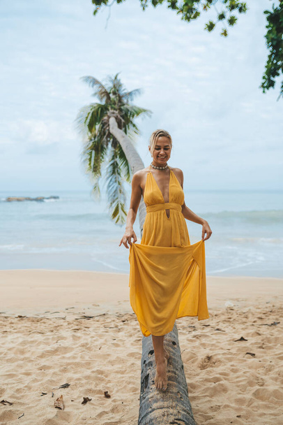 Happy young woman in yellow dress walking along the trunk of a palm tree on the beach - Фото, зображення