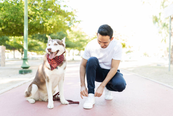 Ισπανόφωνος ιδιοκτήτης σκυλιών που δένει τα κορδόνια του στον ιππόδρομο του πάρκου δίπλα στον σκύλο του. - Φωτογραφία, εικόνα