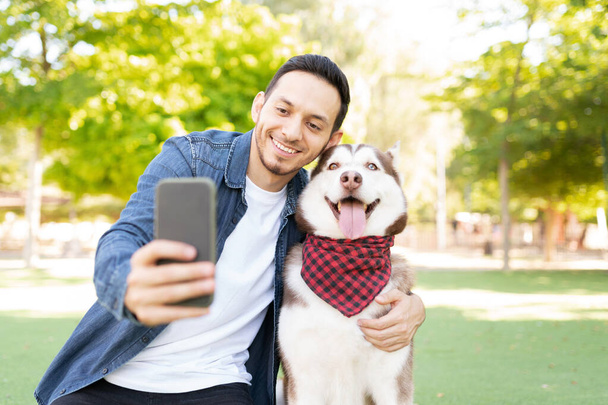 Front view of a good looking man in his 30s taking a photo with his beautiful dog wearing a bandana in the grass at the park - Foto, imagen